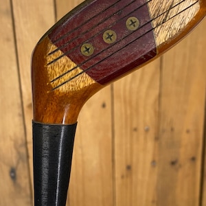 Puede incluir: Primer plano de un palo de golf de madera vintage. La cabeza del palo presenta una combinaci&oacute;n de madera clara y oscura, con una inserci&oacute;n de color burdeos asegurada por tres tornillos de lat&oacute;n. El agarre es negro y texturizado.