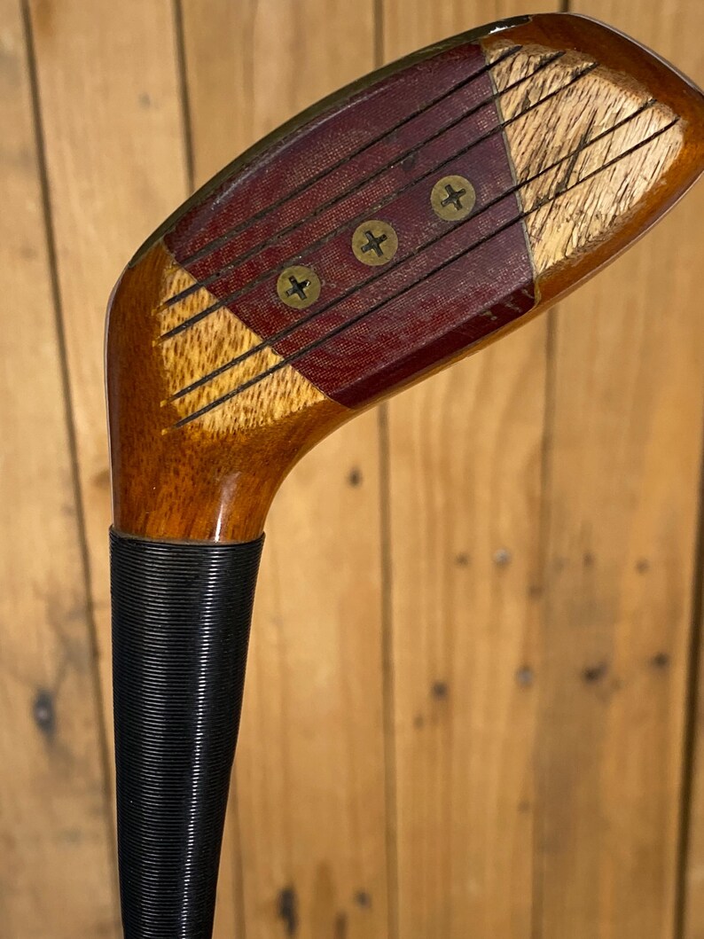 Puede incluir: Primer plano de un palo de golf de madera vintage. La cabeza del palo presenta una combinaci&oacute;n de madera clara y oscura, con una inserci&oacute;n de color burdeos asegurada por tres tornillos de lat&oacute;n. El agarre es negro y texturizado.