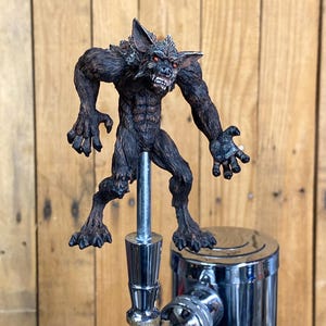 May include: A brown and black werewolf figurine with red eyes, standing on a silver beer tap.