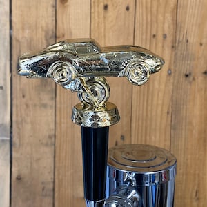 May include: A gold-colored car-shaped beer tap handle with a black handle. The tap handle is mounted on a chrome beer tap.