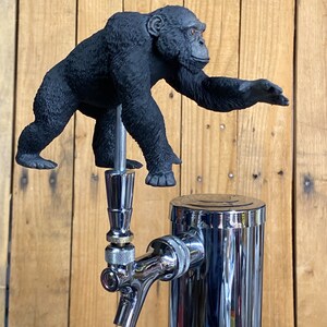 May include: A black gorilla figurine on top of a chrome beer tap. The gorilla is reaching out with its right hand.