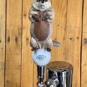 May include: A beer tap handle featuring a sculpted brown and grey groundhog standing upright on top of a white golf ball. The golf ball is printed with the blue and yellow text Keystone Light and the words Always Smooth in smaller blue letters.