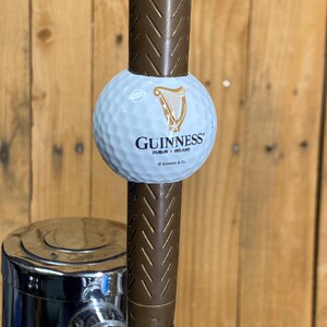 May include: A Guinness-branded golf ball tap handle. The white golf ball features the Guinness logo in gold and black. The handle is brown with a textured design, and it has a chrome base.