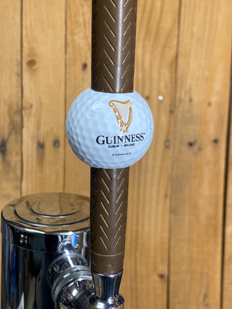 May include: A Guinness-branded golf ball tap handle. The white golf ball features the Guinness logo in gold and black. The handle is brown with a textured design, and it has a chrome base.