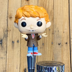 May include: A red-haired figurine with a brown jacket, blue jeans, and red shoes. The figurine is standing on a chrome beer tap.