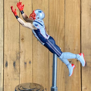 May include: A figurine of an American football player in a diving catch pose. The player wears a silver helmet, red gloves, and a blue and white uniform. The figure is mounted on a chrome tap handle.