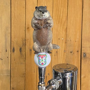 May include: A novelty beer tap handle featuring a groundhog figure atop a golf ball. The groundhog is brown and gray, with a stern expression. The golf ball has a colorful logo. The tap handle is made of polished metal.