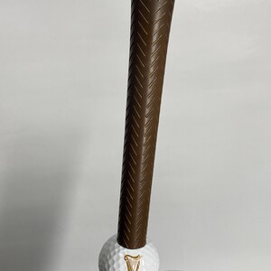 May include: A brown golf club with a textured grip, holding a white golf ball with the Guinness logo. The club is positioned vertically against a plain background. The golf ball is white with the Guinness logo.