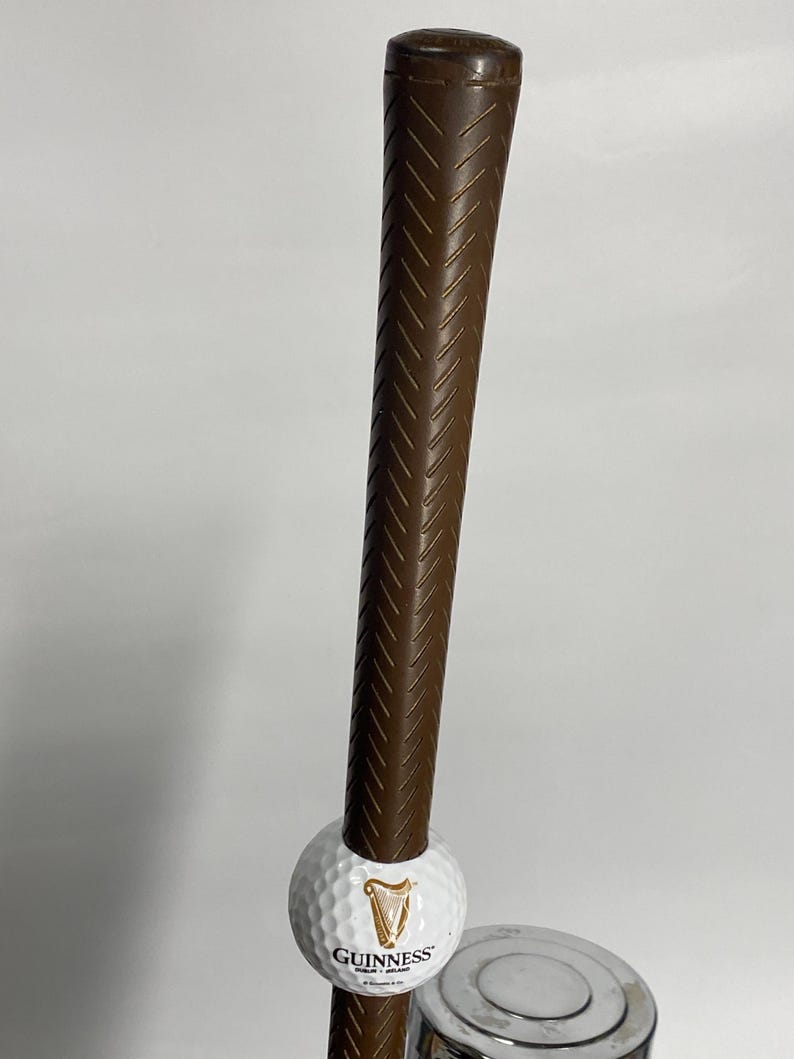 May include: A brown golf club with a textured grip, holding a white golf ball with the Guinness logo. The club is positioned vertically against a plain background. The golf ball is white with the Guinness logo.