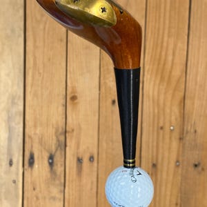 Puede incluir: Un palo de golf antiguo con cabeza de madera y empu&ntilde;adura negra. Una pelota de golf blanca est&aacute; unida al palo. La pelota de golf tiene la palabra "Callaway" impresa. El palo est&aacute; sobre un fondo de madera.