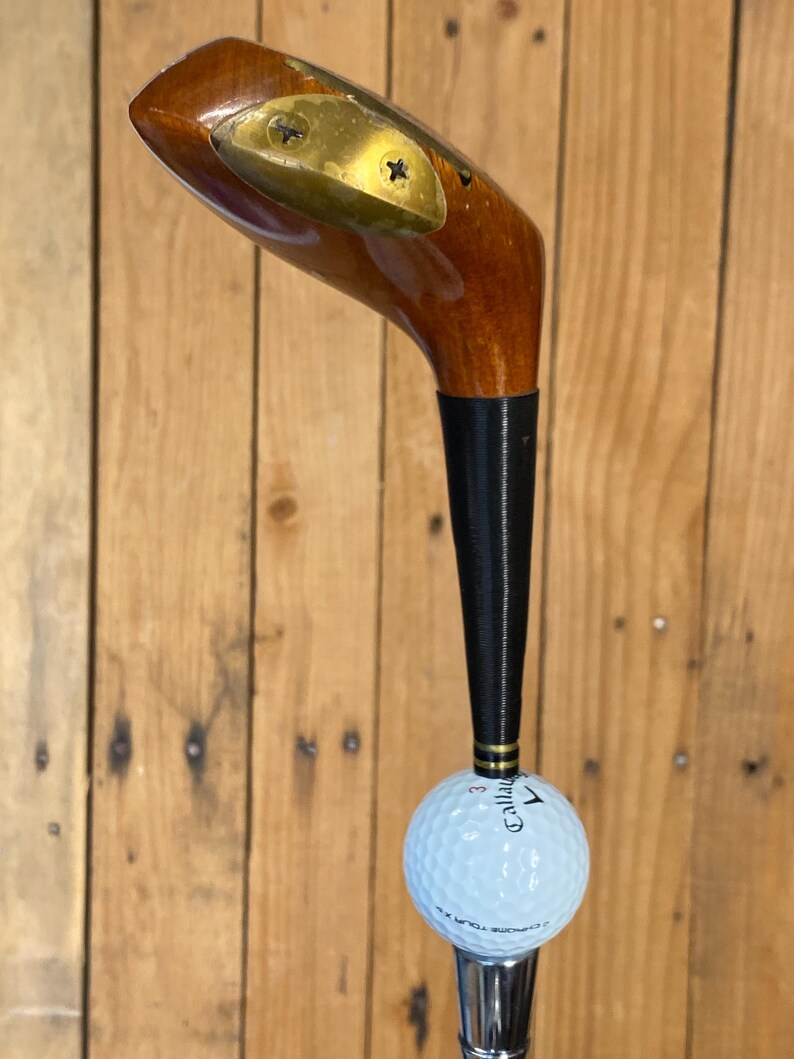 Puede incluir: Un palo de golf antiguo con cabeza de madera y empu&ntilde;adura negra. Una pelota de golf blanca est&aacute; unida al palo. La pelota de golf tiene la palabra "Callaway" impresa. El palo est&aacute; sobre un fondo de madera.