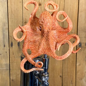 May include: A bright orange octopus beer tap handle with a detailed textured surface. The octopus has eight tentacles that are curled and twisted. The tap handle is attached to a silver metal tap.