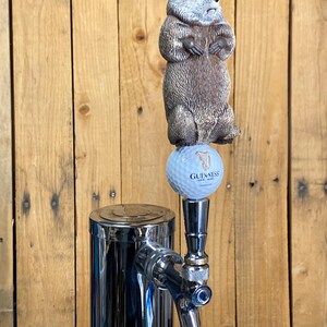 May include: A chrome beer tap with a groundhog figurine on top. The groundhog is brown and gray, standing on a golf ball with the Guinness logo. The tap is set against a wooden background.