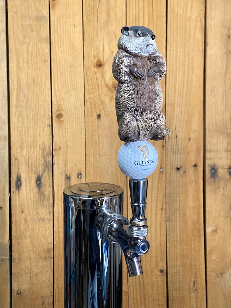 May include: A chrome beer tap with a groundhog figurine on top. The groundhog is brown and gray, standing on a golf ball with the Guinness logo. The tap is set against a wooden background.