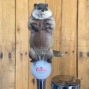 May include: A novelty beer tap handle featuring a groundhog standing on a golf ball. The golf ball has the Coors Light logo. The groundhog is brown and gray with a stern expression. The tap handle is made of metal.