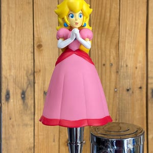 May include: A beer tap handle featuring a molded figure of Princess Peach with blonde hair, a gold crown, blue earrings, white gloves, and a pink dress with a red hem, mounted on a polished chrome metal base with a textured grip section.