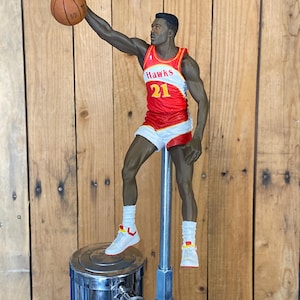 May include: A red, white, and gold Atlanta Hawks basketball player figurine holding a basketball. The figurine is mounted on a silver tap handle.