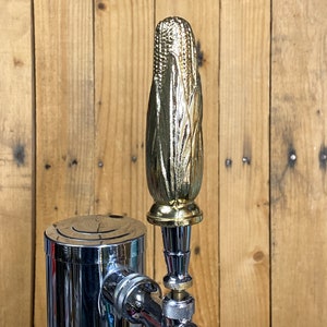 May include: A gold-colored metal beer tap handle shaped like an ear of corn. The handle is attached to a chrome beer tap.