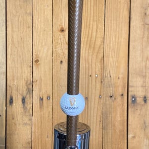 May include: A brown tap handle with a golf club grip design. A light blue golf ball with the Guinness logo is attached. The tap is mounted on a chrome base, set against a wooden background.