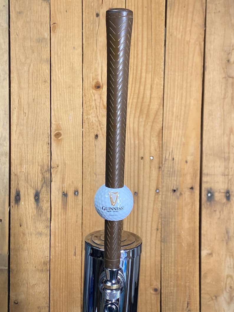 May include: A brown tap handle with a golf club grip design. A light blue golf ball with the Guinness logo is attached. The tap is mounted on a chrome base, set against a wooden background.