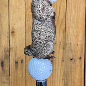 May include: A novelty golf club accessory featuring a detailed groundhog figurine atop a golf ball. The groundhog is brown and gray, with a textured fur appearance. The golf ball is white, and the base is a shiny silver metal.