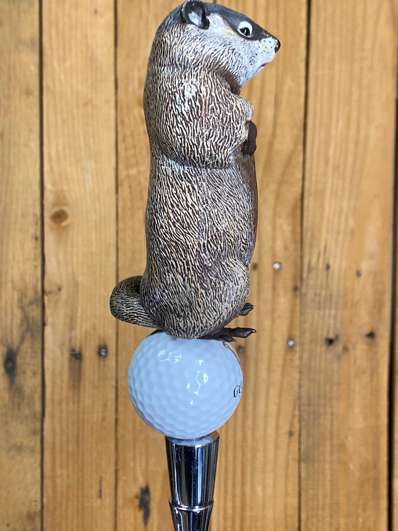 May include: A novelty golf club accessory featuring a detailed groundhog figurine atop a golf ball. The groundhog is brown and gray, with a textured fur appearance. The golf ball is white, and the base is a shiny silver metal.