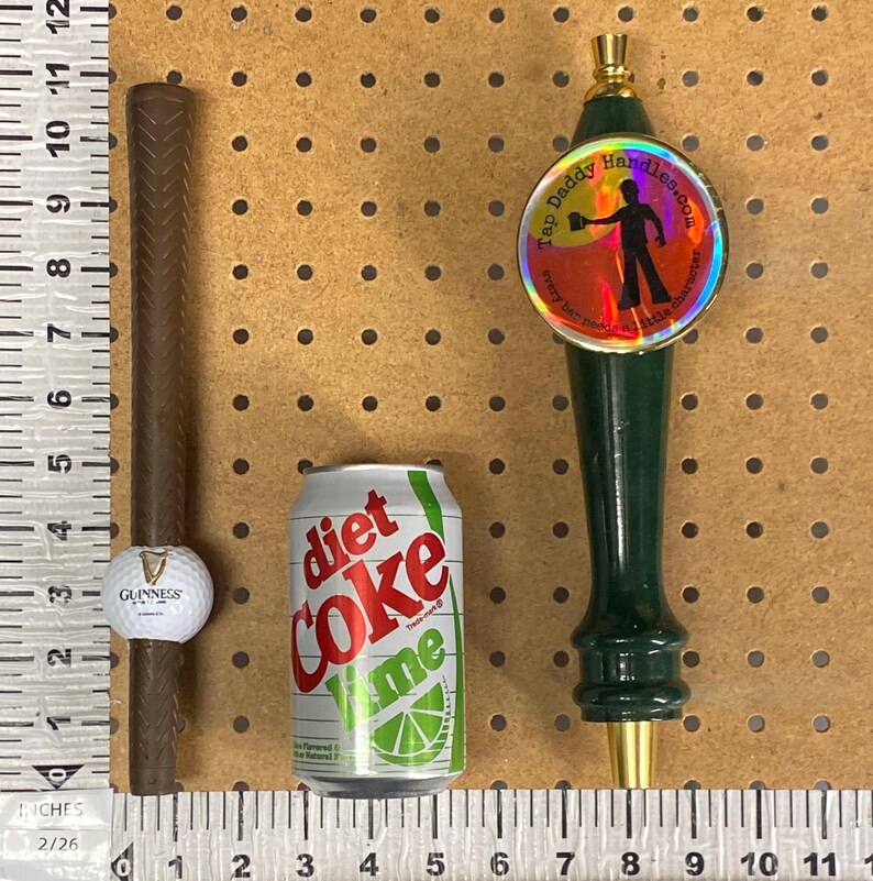 May include: A collection of items: a brown golf club grip with a Guinness golf ball, a Diet Coke Lime can, and a green tap handle with the text "Tap Daddy Handles.com".