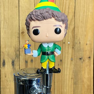 May include: A green and yellow Buddy the Elf Pop figure with a red feather on his hat. He is holding a small toy in his right hand. The figure is attached to a chrome beer tap.