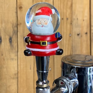 May include: A Christmas-themed tap handle featuring a snow globe with a Santa Claus figure inside. The Santa figure is wearing a red suit and hat, with a white beard and holding a small blue gift. The handle is attached to a chrome tap.