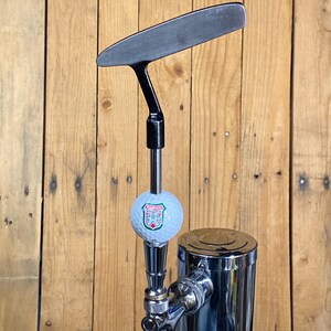 May include: A chrome beer tap with a golf ball-shaped handle and a black putter-shaped spout. The golf ball has a green and red logo on it.