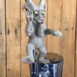 May include: A whimsical, gray creature figurine with large eyes and pointed ears, mounted on a chrome tap handle. The creature is in a waving pose, adding a playful touch. The background is a wooden surface.