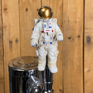 May include: A decorative beer tap handle featuring a small astronaut figure in a white spacesuit with a gold helmet. The astronaut is standing on top of a chrome tap. The background is a wooden surface.