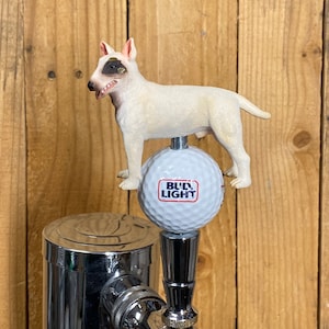 May include: A beer tap handle featuring a white Bull Terrier figurine standing on top of a white golf ball with the Bud Light logo printed in red and blue. The tap handle is constructed from polished chrome metal and is mounted on a vertical base.