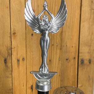 May include: A silver-colored tap handle featuring a winged female figure holding a torch aloft. The figure is detailed with flowing lines and is mounted on a black and silver base. The background is a wooden surface.
