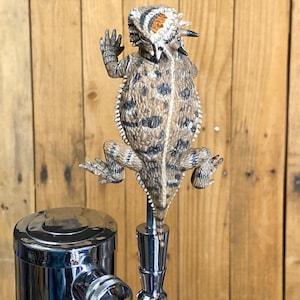 May include: A detailed, hand-painted beer tap handle featuring a horned lizard design. The lizard is primarily brown with black and tan spots, and is mounted on a polished chrome tap. The background is a wooden surface.