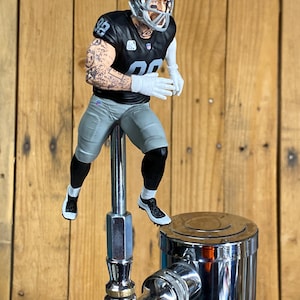 May include: A beer tap handle featuring a miniature football player figurine wearing a black Raiders jersey with the number 98, a silver helmet, grey pants, and white gloves. The player has visible arm tattoos and is mounted on a chrome metal post.