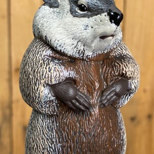 May include: A detailed figurine of a groundhog, featuring a gray and white head, brown fur, and dark brown paws. The groundhog is depicted in a standing pose, with its paws clasped together. The figurine has a textured appearance.