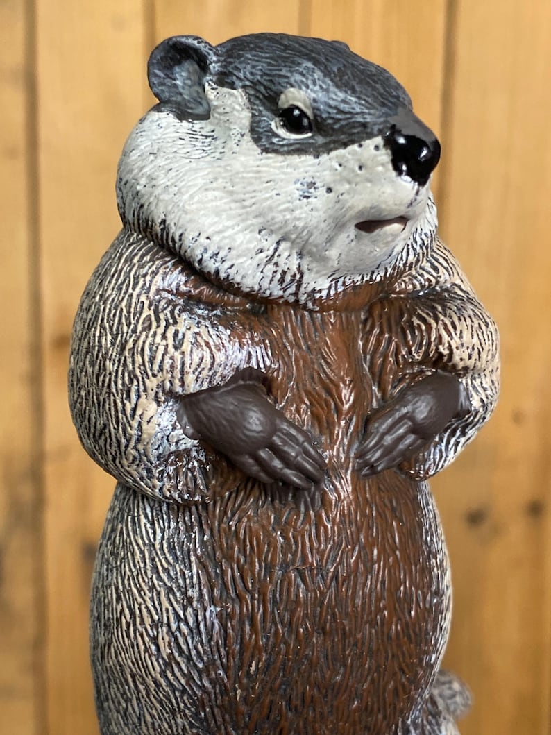 May include: A detailed figurine of a groundhog, featuring a gray and white head, brown fur, and dark brown paws. The groundhog is depicted in a standing pose, with its paws clasped together. The figurine has a textured appearance.