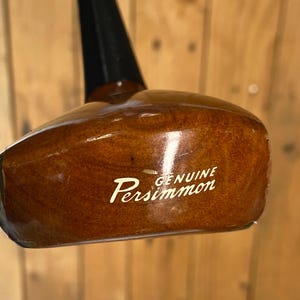 Puede incluir: Una cabeza de palo de golf vintage de madera pulida, con un eje negro. Las palabras "GENUINE Persimmon" est&aacute;n impresas en escritura blanca en la cabeza del palo. La madera tiene un color marr&oacute;n c&aacute;lido y rico.