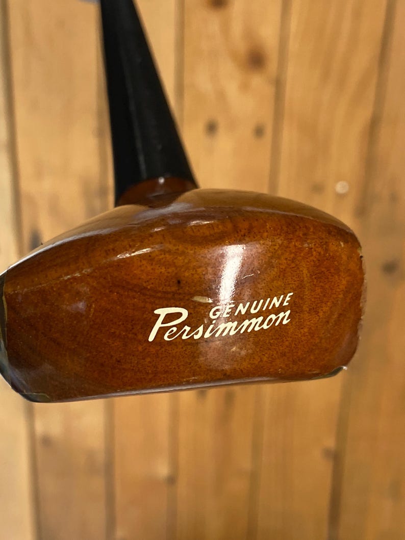 Puede incluir: Una cabeza de palo de golf vintage de madera pulida, con un eje negro. Las palabras "GENUINE Persimmon" est&aacute;n impresas en escritura blanca en la cabeza del palo. La madera tiene un color marr&oacute;n c&aacute;lido y rico.
