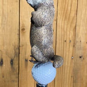 May include: A detailed, hand-painted groundhog figurine atop a golf ball, mounted on a chrome tap handle. The groundhog is brown and gray, with a detailed fur texture. The golf ball is light blue.