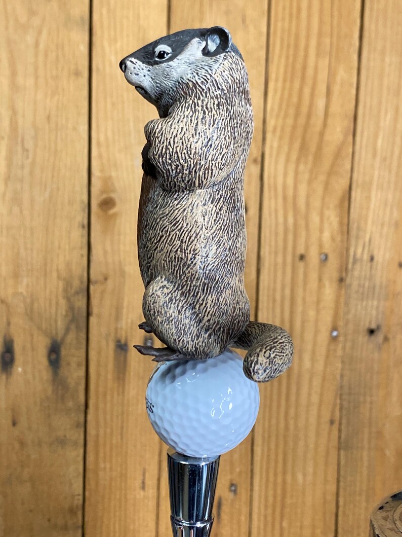 May include: A detailed, hand-painted groundhog figurine atop a golf ball, mounted on a chrome tap handle. The groundhog is brown and gray, with a detailed fur texture. The golf ball is light blue.