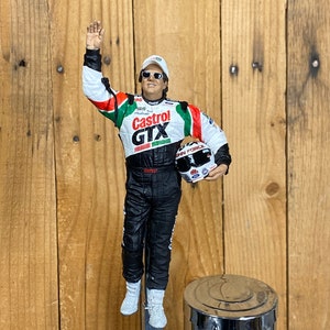 May include: A white, green, and red racing suit with a Castrol GTX logo. The figure is wearing a white cap and sunglasses and is holding a white helmet with a John Force logo. The figure is mounted on a chrome beer tap.
