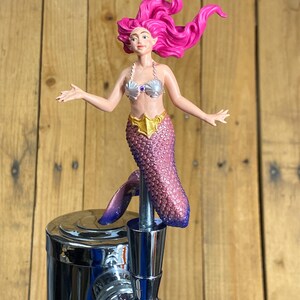 Peut inclure: Une poignée de tireuse à bière surmontée d'une figurine de sirène aux cheveux rose vif, portant un haut en forme de coquillages argentés avec une gemme violette et une ceinture dorée. La queue texturée est irisée, passant du rose au violet.