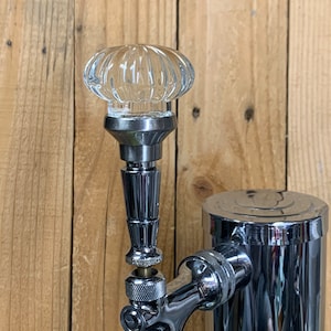 May include: A chrome beer tap with a clear glass knob on top. The tap is attached to a chrome keg.