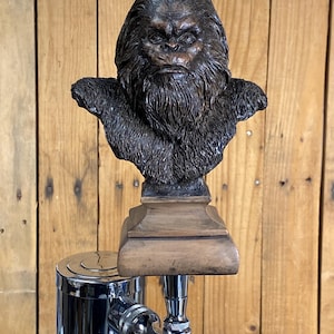 May include: A dark brown, textured bust of a hairy, ape-like creature with a stern facial expression, mounted on a square, light brown wooden base. The bust is attached to a polished, silver-colored metal beer tap handle assembly.