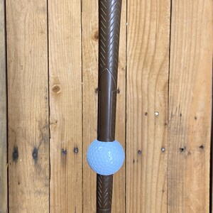 May include: A brown golf club with a light blue golf ball balanced on the club's head. The club features a textured grip and a silver metal base. The backdrop is a wooden wall.