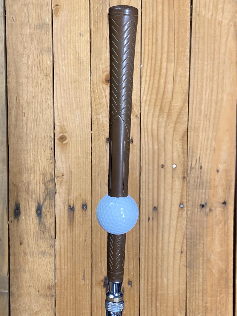 May include: A brown golf club with a light blue golf ball balanced on the club's head. The club features a textured grip and a silver metal base. The backdrop is a wooden wall.