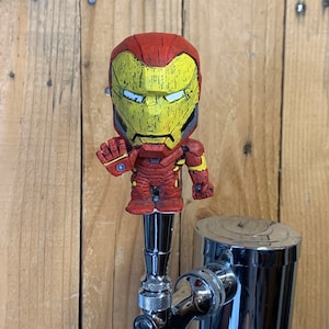 May include: A red and yellow Iron Man tap handle with a chrome faucet. The tap handle features a detailed Iron Man figure with a raised hand. The background is a wooden surface.