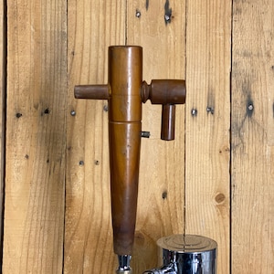 May include: A wooden beer tap with a chrome base. The tap has a wooden handle and a side lever. The tap is mounted on a wooden wall.